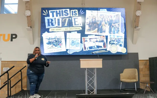 Kayla Life speaking with a microphone on stage in front of a video wall reading This is RITE, our story, why Baltimore