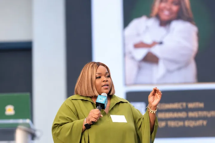 Kayla Life speaking on stage with a microphone in a green outfit, in front of a screen that reads Connect with Rebrand Institute of Tech Equity